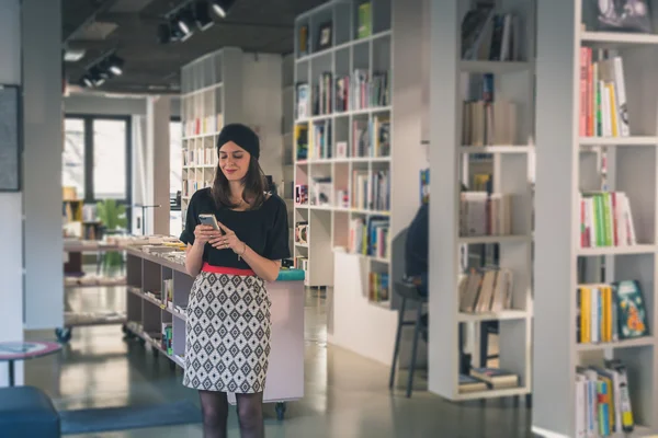 Beautiful young brunette texting in a bookstore — Stock Photo, Image