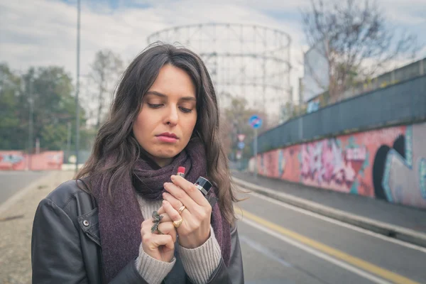 Beautiful young brunette putting on lipstick in the city streets — Stock Photo, Image