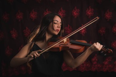 Siyah elbiseli kız, saçları müziğin ritmiyle dans ederken tutkuyla keman çalıyor. Arkasında kırmızı desenli bir arka plan var.