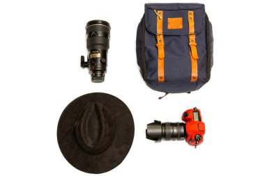 Dağlarda yürüyüş yapmak için basit bir kamp malzemesi. Deri kayışlı mavi bir sırt çantası, kamera, telefoto lensi, beyaz bir arka planda şapka.