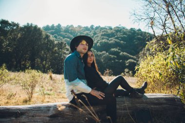 Genç çift, gün batımında ormanda, kuru bir ağaç gövdesinde oturuyor, gülümsüyor ve birlikte vakit geçiriyorlar, adam şapka ve mavi kot gömlek giyiyor ve kız siyah giyiniyor.