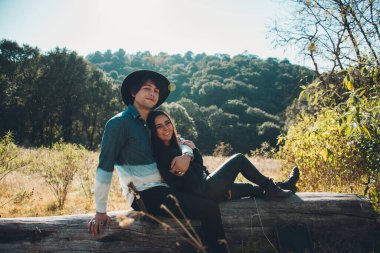 Genç çift, gün batımında ormanda, kuru bir ağaç gövdesinde oturuyor, gülümsüyor ve birlikte vakit geçiriyorlar, adam şapka ve mavi kot gömlek giyiyor ve kız siyah giyiniyor. 7