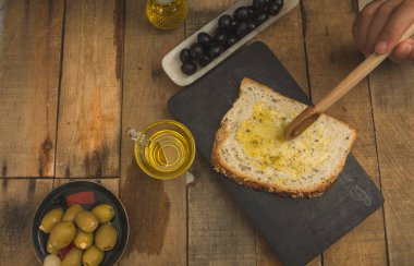 Tahıl ekmeği, dilimler halinde kesilmiş zeytin yağı, kadın eli tarafından ahşap kaşık sürülmüş, doldurulmuş yeşil zeytin, siyah bir tabakta ve ahşap bir arka planda..