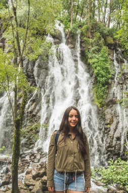 Adventurous girl exploring waterfalls in the middle of the jungle, with light clothes finds amazing landscapes. She wears a green windbreaker and blue denim shorts.