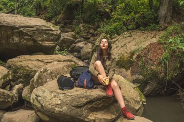Adventurous girl exploring waterfalls in the middle of the jungle, with light clothes finds amazing landscapes. She wears a green windbreaker and blue denim shorts.