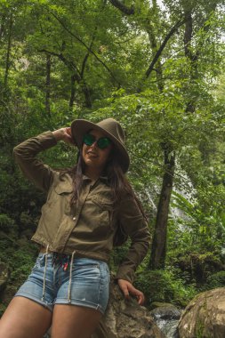 Adventurous girl exploring waterfalls in the middle of the jungle, with light clothes finds amazing landscapes. She wears a green windbreaker and blue denim shorts.