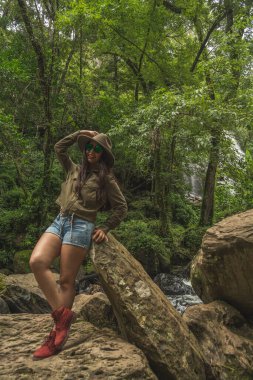 Adventurous girl exploring waterfalls in the middle of the jungle, with light clothes finds amazing landscapes. She wears a green windbreaker and blue denim shorts.