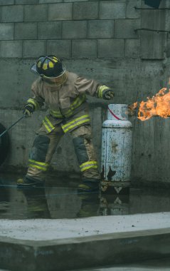 İtfaiyeci kıyafetiyle yanan bir benzin deposunun gaz akışını kahramanca kesiyor.