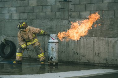 İtfaiyeci kıyafetiyle yanan bir benzin deposunun gaz akışını kahramanca kesiyor.