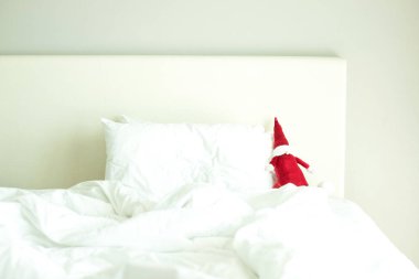 Evde ya da otelde Noel tatili konsepti, rahat yatak odası, yalnızlık, huzur. Temiz beyaz çarşafları, yumuşak yastıkları ve battaniyesi olan büyük boş bir yatakta şirin kırmızı Noel Baba oyuncağı. Yatay olarak. Boşluğu kopyala. 