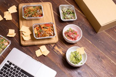 Çevrimiçi sipariş, teslimat, sağlıklı ve temiz yemek kavramı. Laptop ve kutunun yakınındaki ahşap arka planda hazır yemek kutularında çeşitli vejetaryen yemekleri. Üst Manzara. Hiç kimse. Tayland eriştesi, vegan humusu..