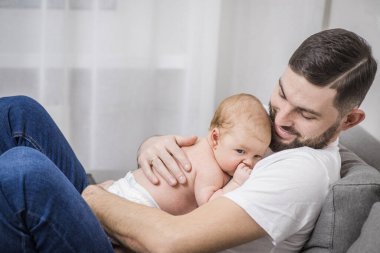 Mutlu, sakallı, sevgi dolu bir baba kucağında yeni doğmuş bir bebek tutuyor. Evdeki kanepede oturuyor. Baba tenli bir bebek. Kafkasyalı, ebeveynlik kavramı, babalık, bir çocuğun hayatının ilk ayları.