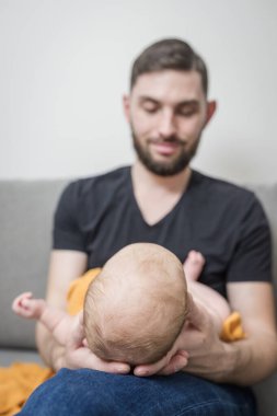 Sakallı sevecen baba yeni doğmuş bebeği sarı muslin bezine sarmış tutuyor. Evdeki kanepede oturuyor. Bebek bakımı konsepti.