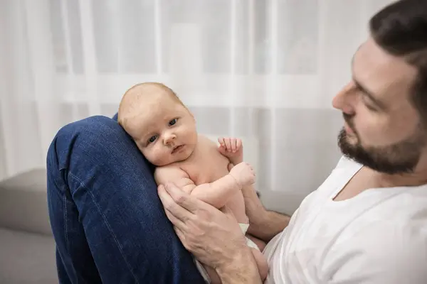 Loving happy father with beard holds newborn baby in his arms. Sits on couch at home. Concept of parenting, paternity, the first months of child\'s life.