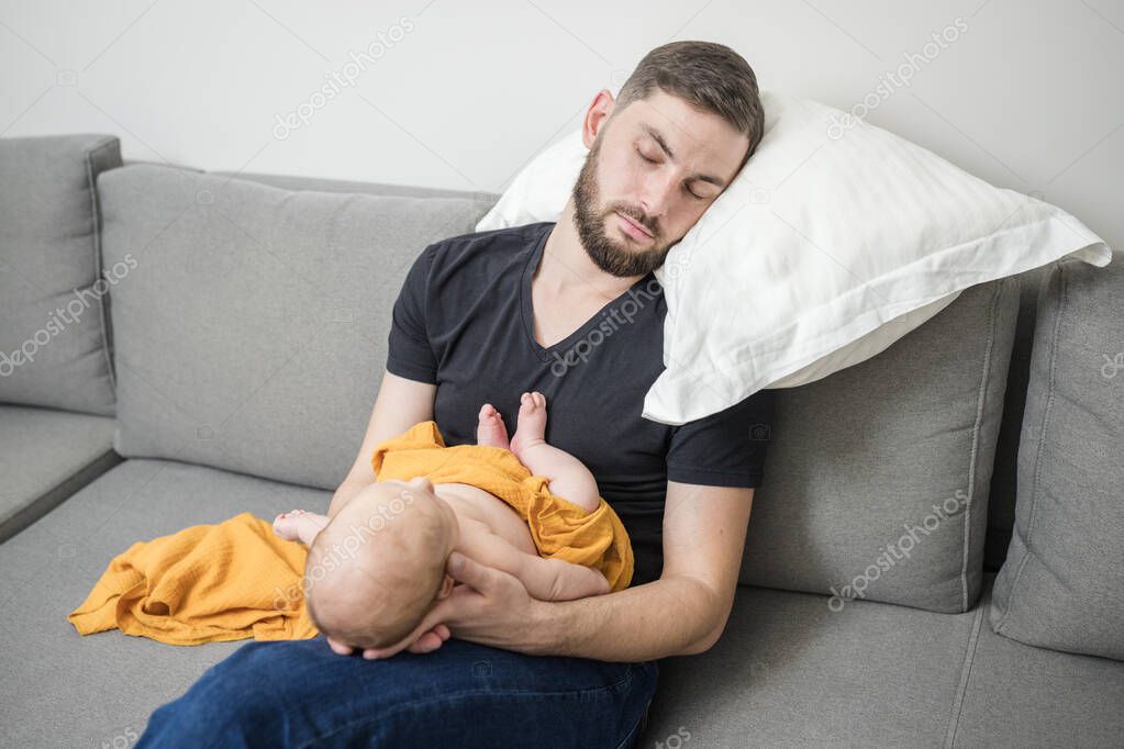 Tired man with beard sleeps while sitting, holding newborn baby in his arms, covered with a trendy yellow muslin diaper. Lifestyle. Concept of lack of normal sleep in first months of newborn's life.