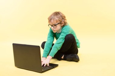 Gözlüklü sarışın liseli çocuk oynuyordu. Laptopta uzaktan ders çalışmak, sarı arka planda bacak bacak üstüne atmak. Uzaklık öğrenimi, IT - eğitim, görme sorunları, ekran zaman kavramı. 