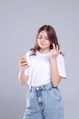 Akıllı telefonlu hoş bir kız telefonuna bakıyor. Stüdyo ışığı, gri arka plan. Kafkasyalı. Beyaz tişört ve kot pantolon giymişti. Gençler için sosyal ağlar, aşk, online ilişkiler