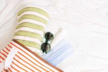 COVID 19 tecridinden sonra seyahat konsepti. Yeni güvenlik kuralları, toplum içinde maske takmak. Plaj eşyaları: tıbbi maske, güneş şapkası, plaj çantası, havlu, güneş gözlüğü oteldeki yatakta beyaz çarşaf arkaplamasına karşı..