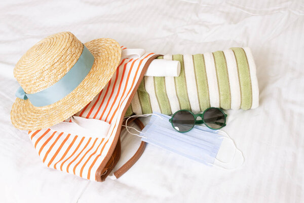 Travel concept after COVID-19 lockdown. New safety rules, wearing masks in public. Beach items: Medical mask, sun hat, beach bag, towel, sunglasses against the background of white linen on the bed in the hotel.