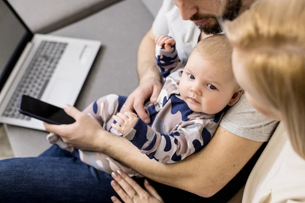 Father working with laptop and mobile phone, looking after his baby. Top view of parents and newborn. Concept of shopping online, home office, work from home, home healthcare, telehealth.