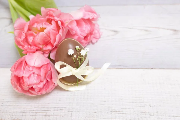 Easter decor, christian religious traditions concept. Beautiful pink tulips, spring flowers, Easter chocolate eggs on background of white boards. Copy space.