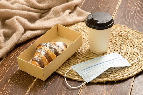 Concept of cafe, bistro during pandemic, takeaway coffee, snack outside home in quarantine, eco friendly food container. Disposable glass with drink, croissant in craft box, face mask on wooden table.