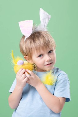 Küçük tatlı duygusal çocuk nazikçe yumurta ve tavşan kulaklı dekoratif yuvayı tutuyor. Yeşil stüdyo geçmişi. Kopya alanı olan Paskalya kartı. Paskalya geleneği, yumurta avcısı kavramı. Göz teması. 