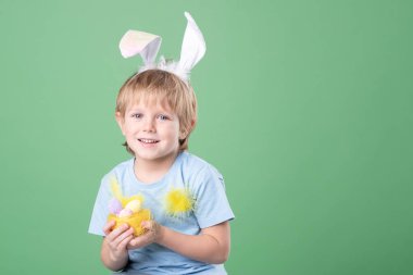 Paskalya yumurtaları ile dekoratif yuvası olan sevimli bir çocuk, ellerinde tüyler, kafasında renkli yeşil arka planda tavşan kulakları. Paskalya kartı, kopyalama alanı. Kutlamaya hazırlanıyor, Paskalya gelenekleri.