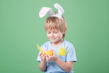 Küçük tatlı duygusal sarışın çocuk nazikçe dekoratif yuvayı yumurtalarla, tavşan kulaklarıyla tutuyor. Yeşil stüdyo geçmişi. Kopya alanı olan Paskalya kartı. Paskalya geleneği, yumurta avcısı kavramı. Yatay