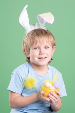 Paskalya yumurtaları ile dekoratif yuvası olan sevimli bir çocuk, ellerinde tüyler, kafasında renkli yeşil arka planda tavşan kulakları. Paskalya kartı, kopyalama alanı. Kutlamaya hazırlanıyor, Paskalya gelenekleri.