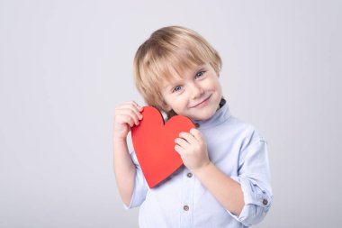 Cute Caucasian child is tenderly holding toy red heart on white background. Eye contact. Studio light. Concept of childhood, love, mother's day, health care, valentine. Horizontal. Copy space.