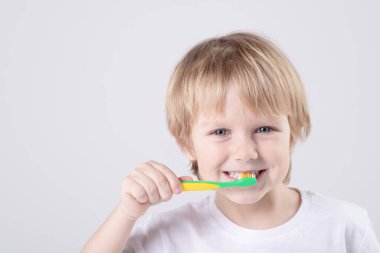 Funny Caucasian child brushes teeth. Eye contact. Studio light. White T-shirt. Concept of baby teeth, healthy lifestyle, routine, development of a child's skill to brush his own teeth. Copy space