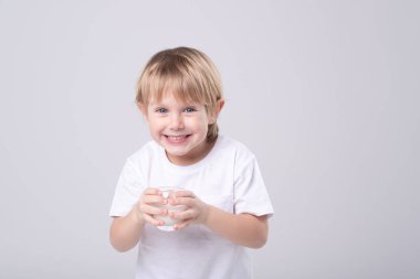 Portrait of a blond Caucasian child with a glass of milk on a white studio background. Expressive face, dental smile, eye contact. Vitamin calcium, milk teeth, dairy products, healthy food concept.