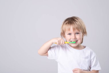 Funny Caucasian child brushes teeth. Eye contact. Studio light. White T-shirt. Concept of baby teeth, healthy lifestyle, routine, development of a child's skill to brush his own teeth. Copy space