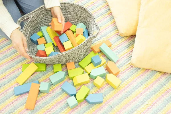 Caucasian woman sits on a striped carpet in a kid\'s room, puts beautiful wooden toys in a basket. Concept of motherhood, cleaning up, toys from eco materials, cozy home. Lifestyle. Horizontal.