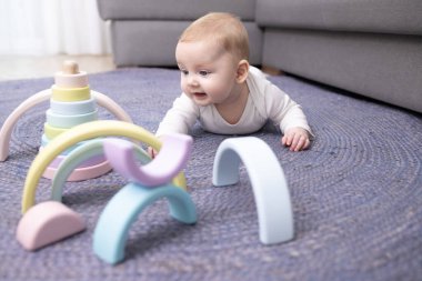 3 aylık beyaz bir bebek emeklemeye çalışıyor. Sevinçle ahşap renkli güzel oyuncaklara bakıyor. Bebek, kanepenin yanındaki mavi hasır halıya yüzüstü uzanıyor. Çocuk gelişimi, sıcak zemin konsepti.