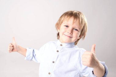 Pozitif gülümseyen bir çocuk başparmak hareketi yaptı. Kafkasyalı, sarı saçlı. Gri bir stüdyoda neşeli, neşeli bir çocuk. Portre. 5-6 yaşlarında bir çocuk..
