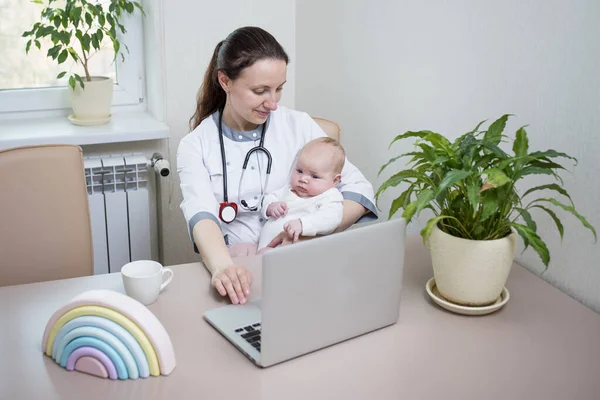 Woman in a medical gown with newborn at the table near laptop. Mom doctor with baby in her arms works online. Topic of online consultation, the work of a doctor from home via the Internet, motherhood.