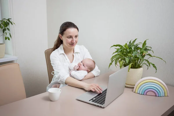 Mom with a newborn baby is working from home, typing on a laptop keyboard in the kitchen. Concept for online work, online shopping, busy mom, online consultation, new life, mother on maternity leave.