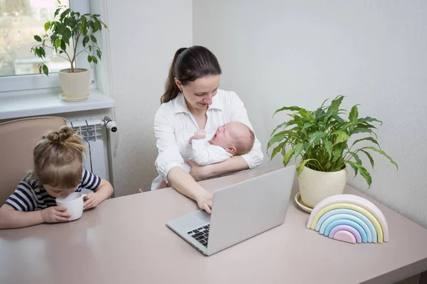 Busy mom with two small children works online at home on laptop. Sitting in kitchen. Crying newborn baby in mother\'s arms, three-year-old girl next to her is drinking from cup. lifestyle family moment