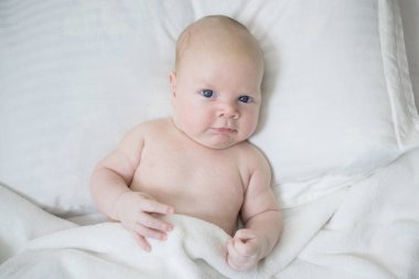 Olumlu bir ifadeyle çıplak yeni doğmuş bebek. Yatakta beyaz bir battaniyeyle kaplanmış bir yastığın üzerinde yatıyor. Beyaz, tombul, şirin bir bebek. Göz teması. Çocuk sahibi olmanın keyfi, sağlıklı bir bebek, günaydın..