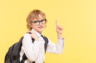 Sarı arka planda izole edilmiş sırt çantalı duygusal okul çocuğunun stüdyo portresi. Gözlüklü mutlu çocuk boş bir yeri işaret ediyor. Reklam konsepti harika fikir, görme, okula dönüş.