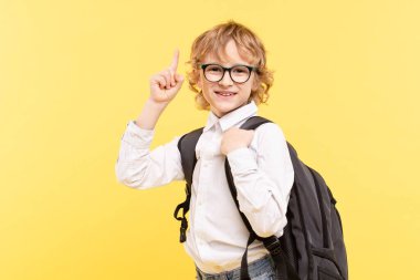 Sarı arka planda izole edilmiş sırt çantalı duygusal okul çocuğunun stüdyo portresi. Gözlüklü mutlu çocuk boş bir yeri işaret ediyor. Reklam konsepti harika fikir, görme, okula dönüş.