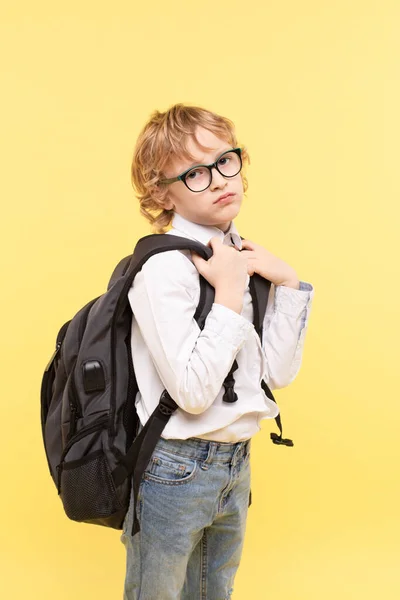 眼鏡をかけた小学校の少年は 大きな重いバックパック 顔の表情を退屈に 白人の男の子 黄色の背景のブロンド 学校に戻るという概念 学校に関する問題 学ぶ意欲のなさ — ストック写真