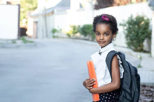 オレンジ色のフォルダを保持バックパックを持つ幸せな学齢の子供 陽気な混合レースの女の子の肖像画 アフロヘア 屋外だ 教育の概念に戻るコピースペース — ストック写真