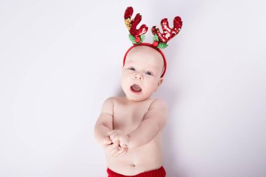 Noel kostümü giymiş mutlu yeni doğmuş bebek beyaz arka planda yatıyor. Stüdyo ışığı. Kafasında kırmızı geyik boynuzları ve aksesuarları var. Komik çocuklar, kış, yeni yıl konsepti. Beyaz bir çocuk. Boşluğu kopyala.