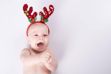 Noel kostümü giymiş mutlu yeni doğmuş bebek beyaz arka planda yatıyor. Stüdyo ışığı. Kafasında kırmızı geyik boynuzları ve aksesuarları var. Komik çocuklar, kış, yeni yıl konsepti. Beyaz bir çocuk. Boşluğu kopyala.