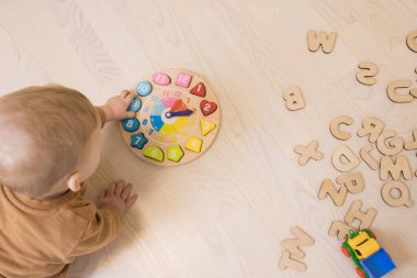 Toddler tahta bir saatle oynuyor, tahta zeminde yatıyor, İngiliz alfabesinin harflerinin yanında. Eğitim oyunları kavramı, erken bebek gelişimi, sağlıklı tahta oyuncaklar için güvenli.