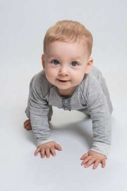 Beyaz stüdyo arka planında sürünen şirin bir bebek, sana bakıyor, gülümsüyor. Çocuk gri tulum giymiş. Mavi gözlü sarışın beyaz çocuk neşeyle gülümsüyor. Göz teması. Bebek reklamı konsepti.