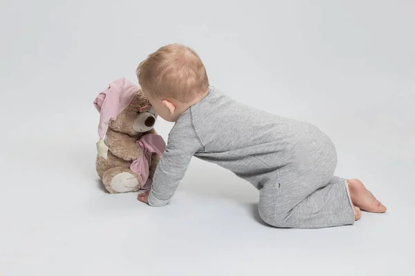 穿着灰色衣服的小孩爬到一只柔软的泰迪熊面前摸了摸 后视镜白色工作室背景 童年概念 6个月至1岁的儿童发育 小宝宝 最喜欢的玩具 图库图片